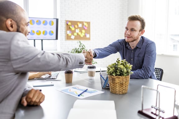 A collaborative meeting concludes with a handshake between two professionals in a modern office, surrounded by charts and work materials.