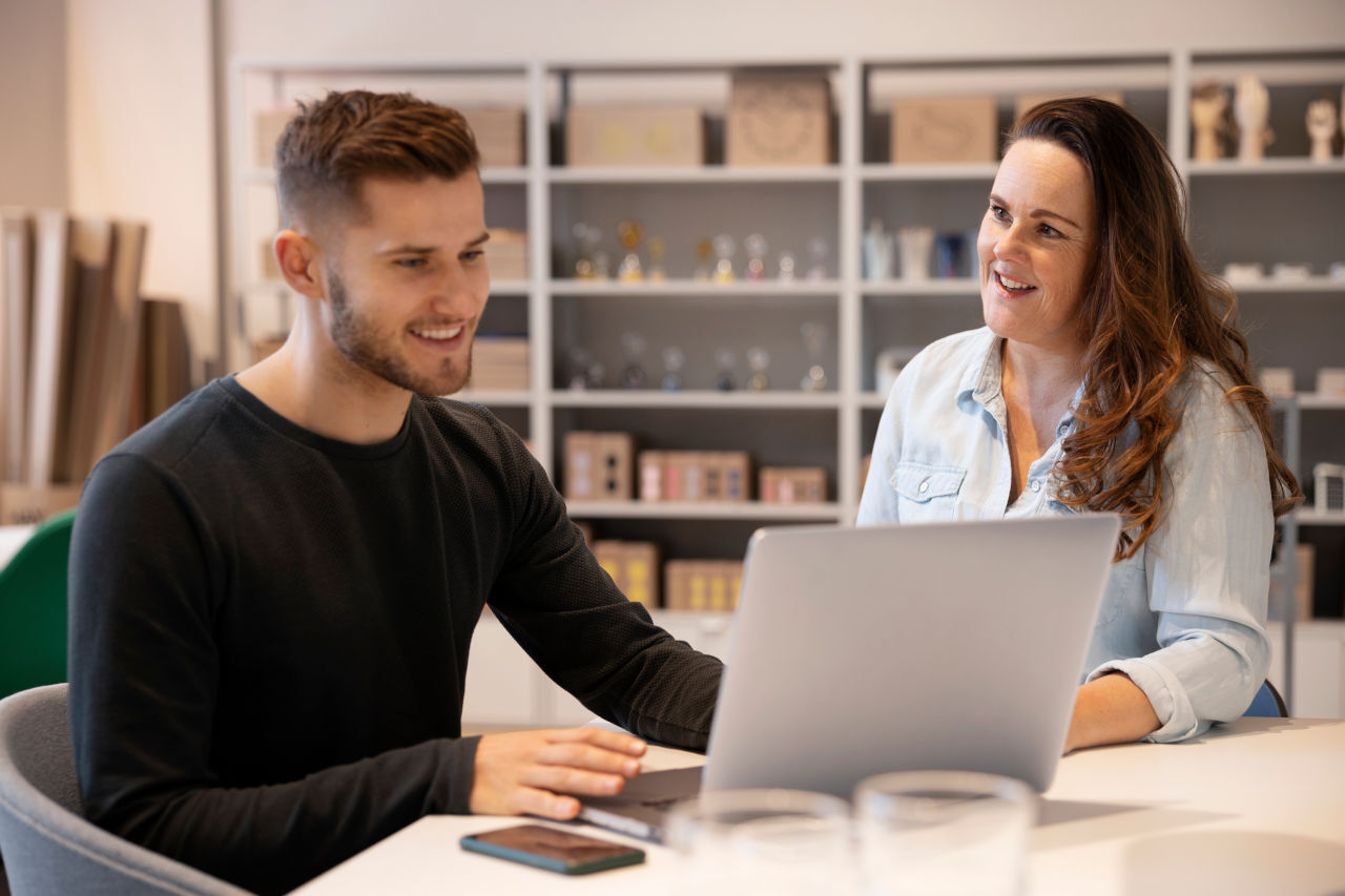Two people sit together at a table, smiling as they look at a laptop in a bright, modern workspace. A smartphone rests on the table, and shelves filled with boxes and decorative items are visible in the softly blurred background.